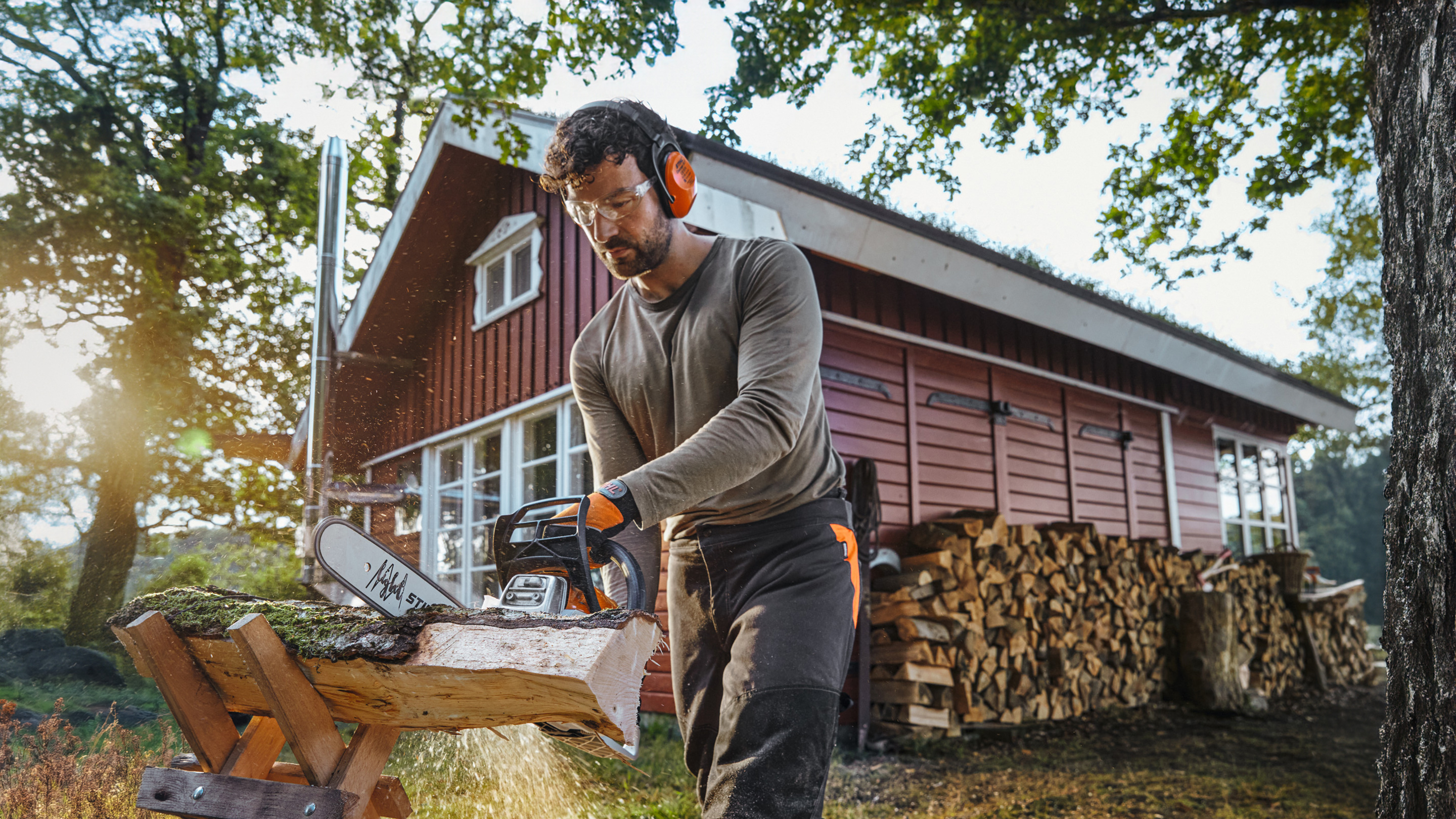 Tronçonneuse STIHL avec laquelle un homme scie un tronc devant une cabane en bois rouge avec une pile de bois de chauffage