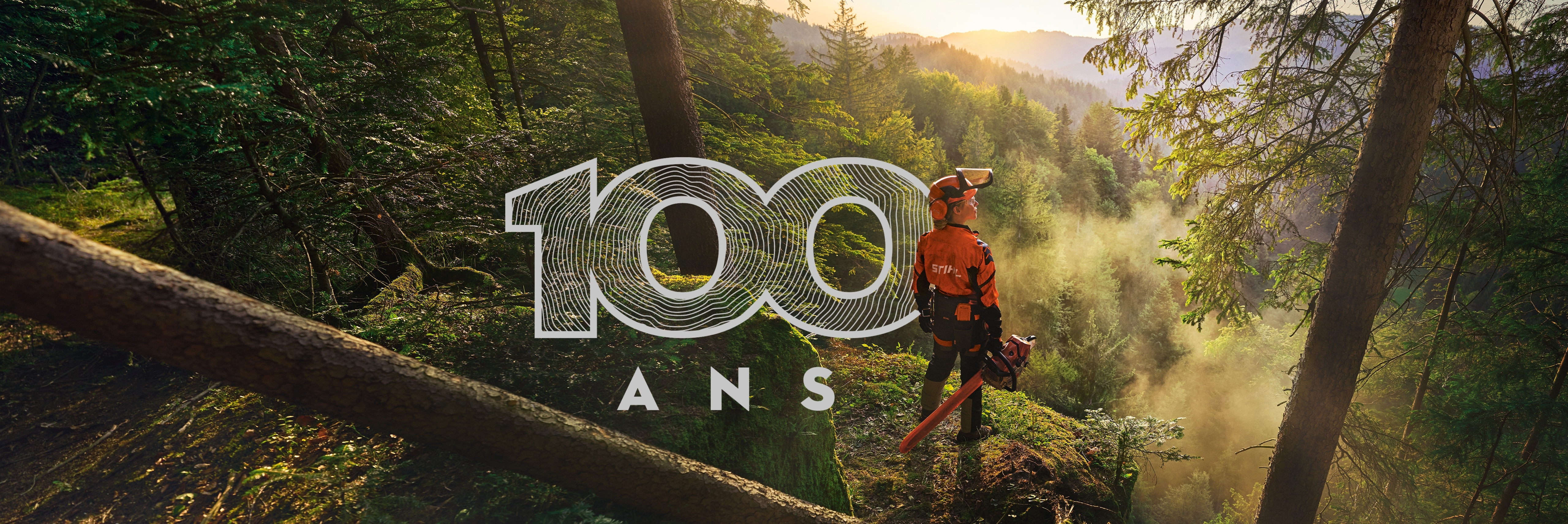 Une femme se tient au bord d'une falaise dans une forêt d'un vert saturé. Elle porte des vêtements de travail STIHL orange, le casque ADVANCE ProCom et tient une tronçonneuse STIHL MS.