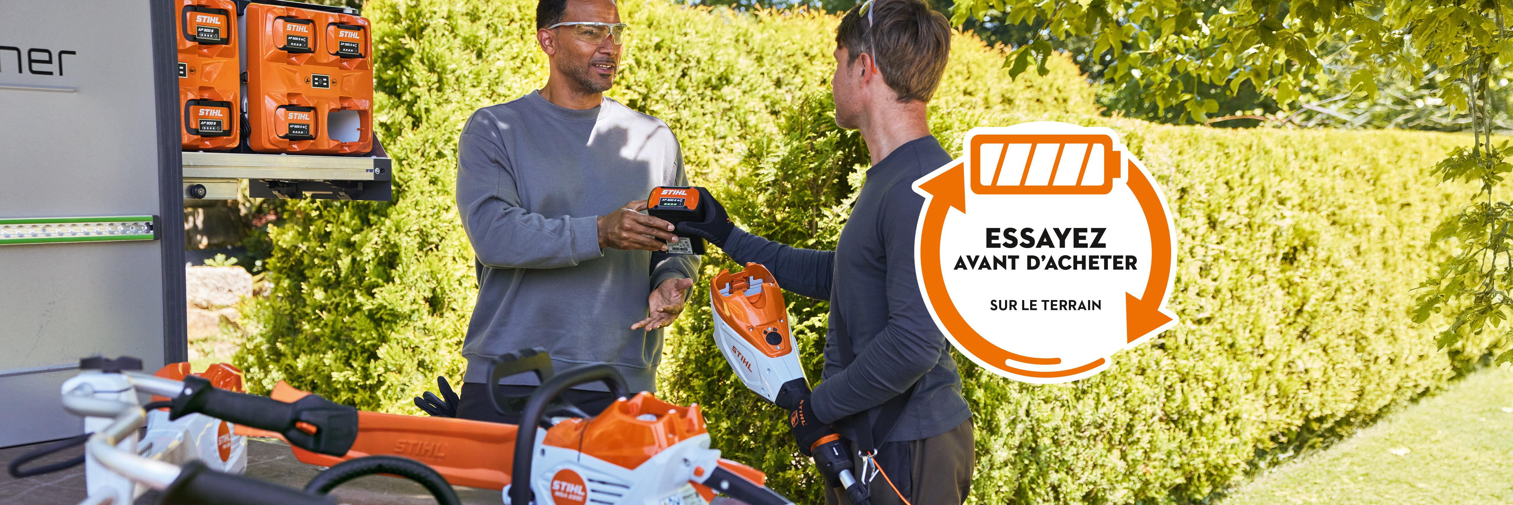 Deux hommes insérant une AP 300 S dans un taille-haies à batterie. Autocollant « Try before Buy » avec l’invitation « Test it Now – contact us for details »